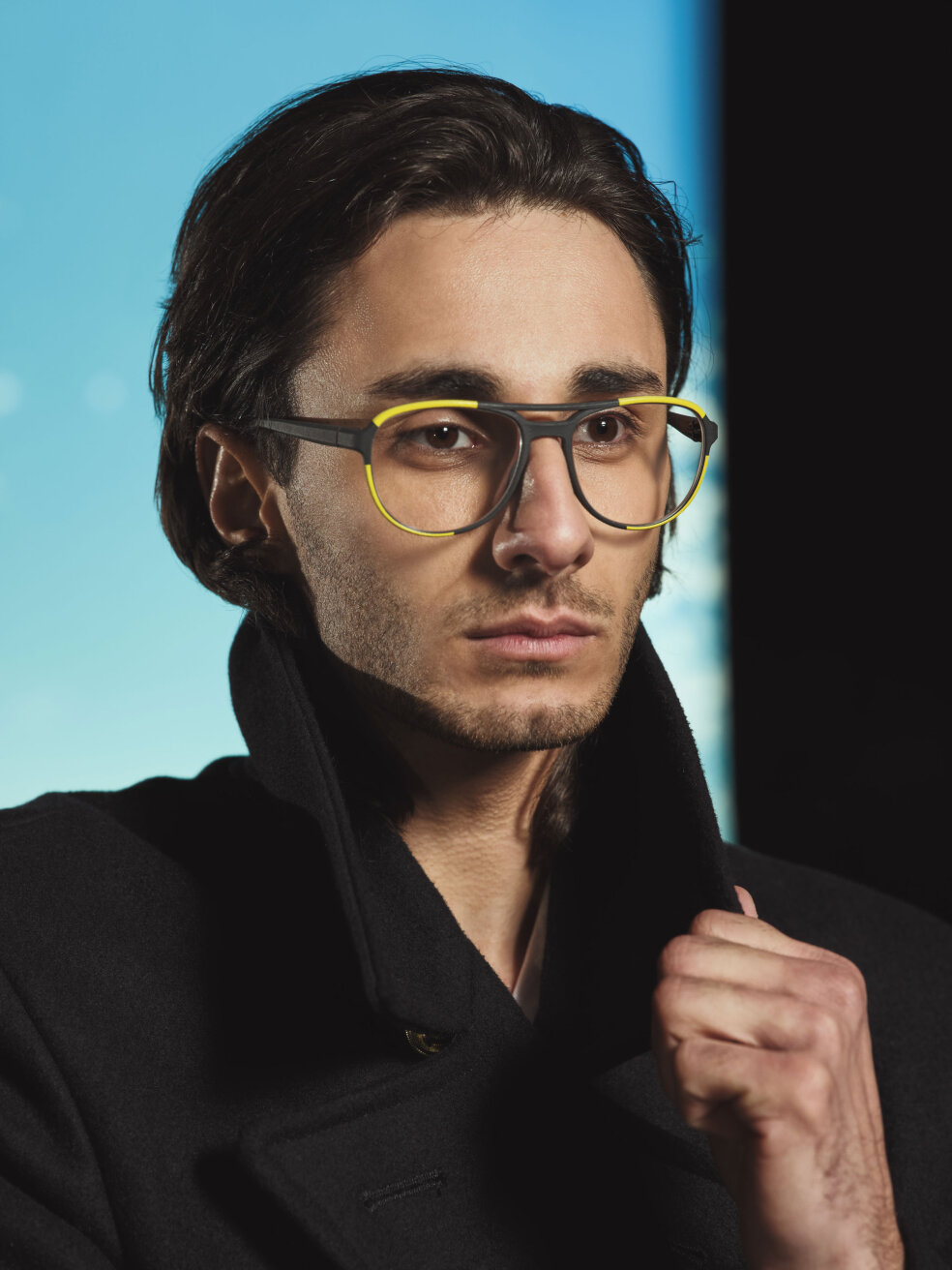A young man with medium-length, dark hair is wearing large, fashionable glasses with yellow rims and a black coat with the collar turned up. The background has a color gradient of blue and black.