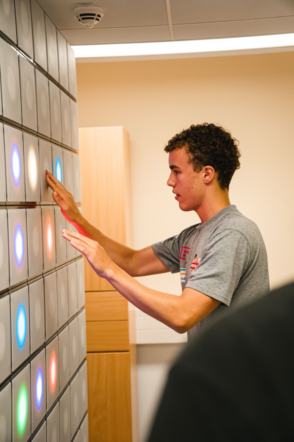 Ein junger Mensch in einem grauen T-Shirt interagiert mit einer Wand aus beleuchteten, bunten Kreisen, möglicherweise als Teil eines interaktiven Spiels oder einer Übung in einer modernen Innenumgebung.