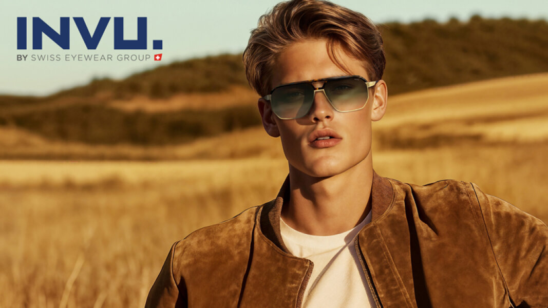 Ein junger Mann mit modischer Sonnenbrille und einer braunen Wildlederjacke steht auf einem sonnenbeschienenen Feld mit H&uuml;geln im Hintergrund. Das Logo der INVU. BY SWISS EYEWEAR GROUP ist in der oberen linken Ecke zu sehen.