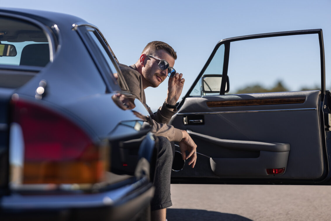 A man with sunglasses sits in a vintage car with the door open and looks into the camera. The shiny exterior of the car and the clear sky suggest a sunny day.