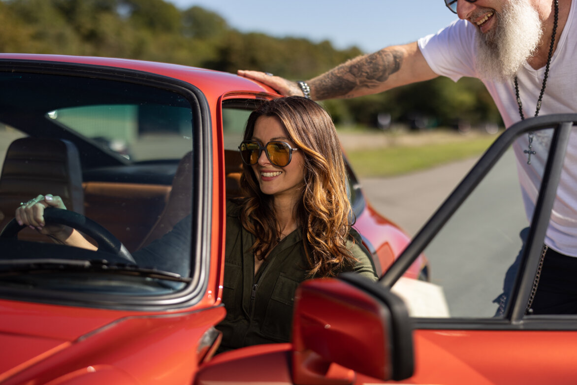 A smiling woman with sunglasses sits in the driver's seat of a red car, while a man with a white beard and sunglasses stands outside leaning against the car door. The background is a green outdoor area.