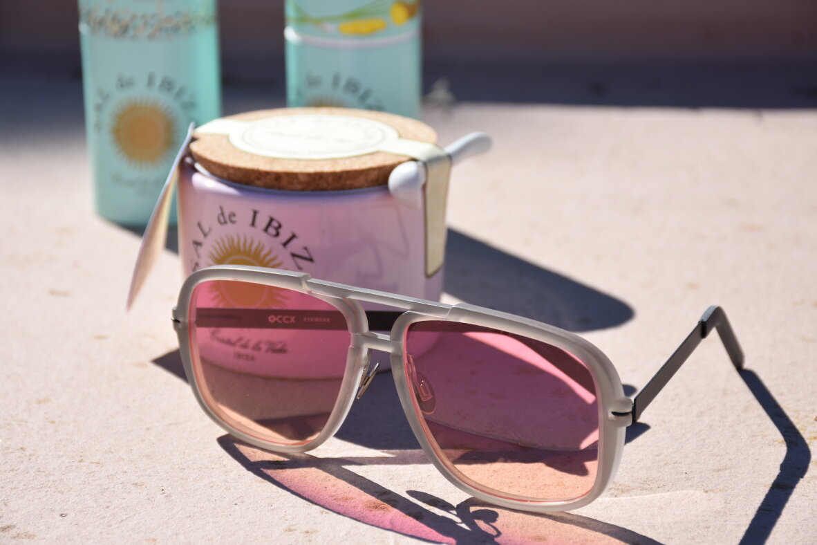 Eine Sonnenbrille mit rosafarbenen Gläsern liegt auf einer hellen Fläche vor einem Glas und einem zylindrischen Behälter mit der Aufschrift "Sal de Ibiza". Die Szene ist durch das Sonnenlicht gut beleuchtet und wirft klare Schatten.