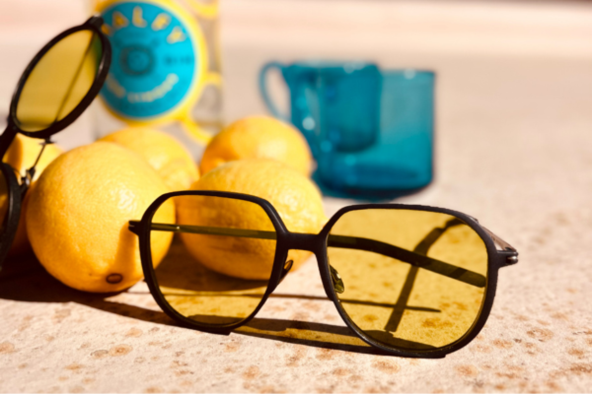 Eine gelb getönte Sonnenbrille liegt auf einer Steinfläche neben Zitronen, einem blauen Becher und einer teilweise sichtbaren gelben Dose im Hintergrund. Helles Sonnenlicht wirft Schatten auf die Szene.