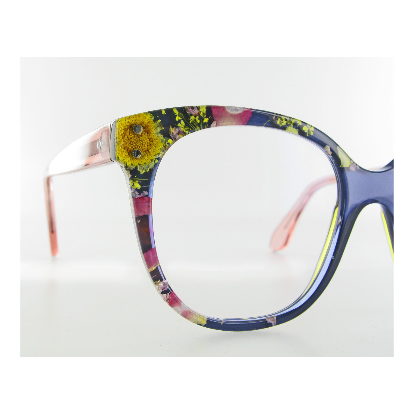 Nahaufnahme einer Brille mit transparentem Rahmen, in den bunte getrocknete Blumen, darunter gelbe und rosa Blüten, vor einem schlichten weißen Hintergrund eingearbeitet sind.
