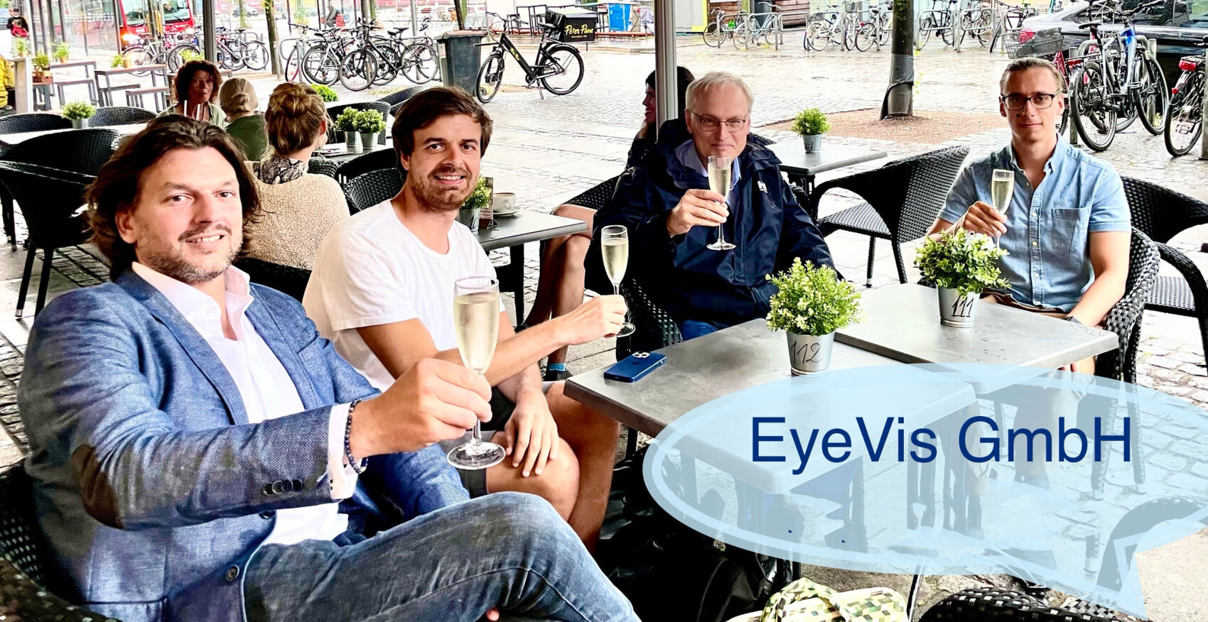 Vier Männer sitzen an einem Cafétisch im Freien, lächeln und erheben ihre Gläser zu einem Toast. Kleine Topfpflanzen schmücken den Tisch. Der Text "EyeVis GmbH" befindet sich auf einem transparenten Overlay in der unteren rechten Ecke. Im Hintergrund sind Fahrräder zu sehen.