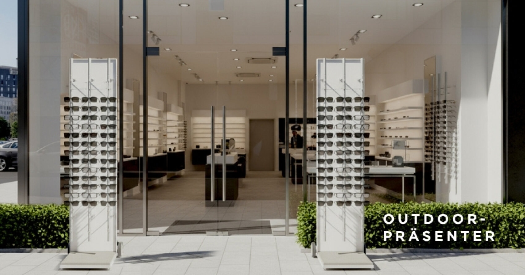 Ein modernes Brillengeschäft mit großen Glasfenstern und zwei Außenständern, die mit Sonnenbrillen gefüllt sind. Im Inneren säumen Brillenregale die Wände. Auf der rechten Seite ist der Schriftzug OUTDOOR-PRÄSENTER zu sehen.