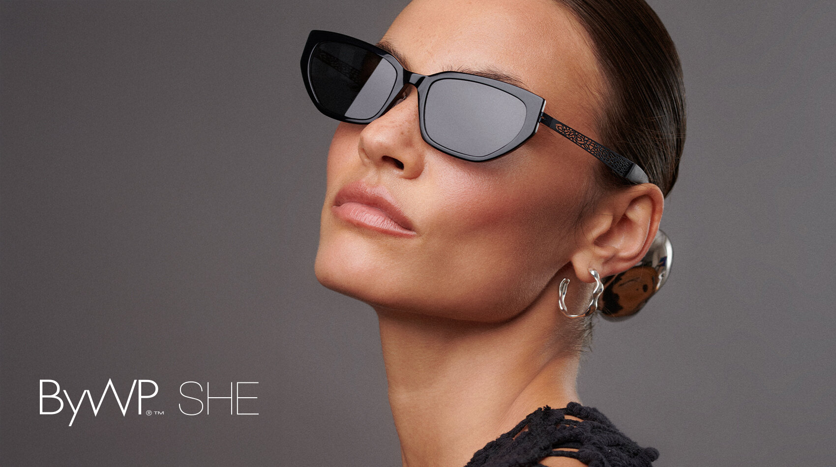 Eine Frau mit zurückgekämmtem Haar trägt eine schwarze Sonnenbrille mit Katzenaugen, Ohrringe und ein schwarzes strukturiertes Oberteil. Der Hintergrund ist grau, und der Text ByWP SHE erscheint in der unteren linken Ecke.