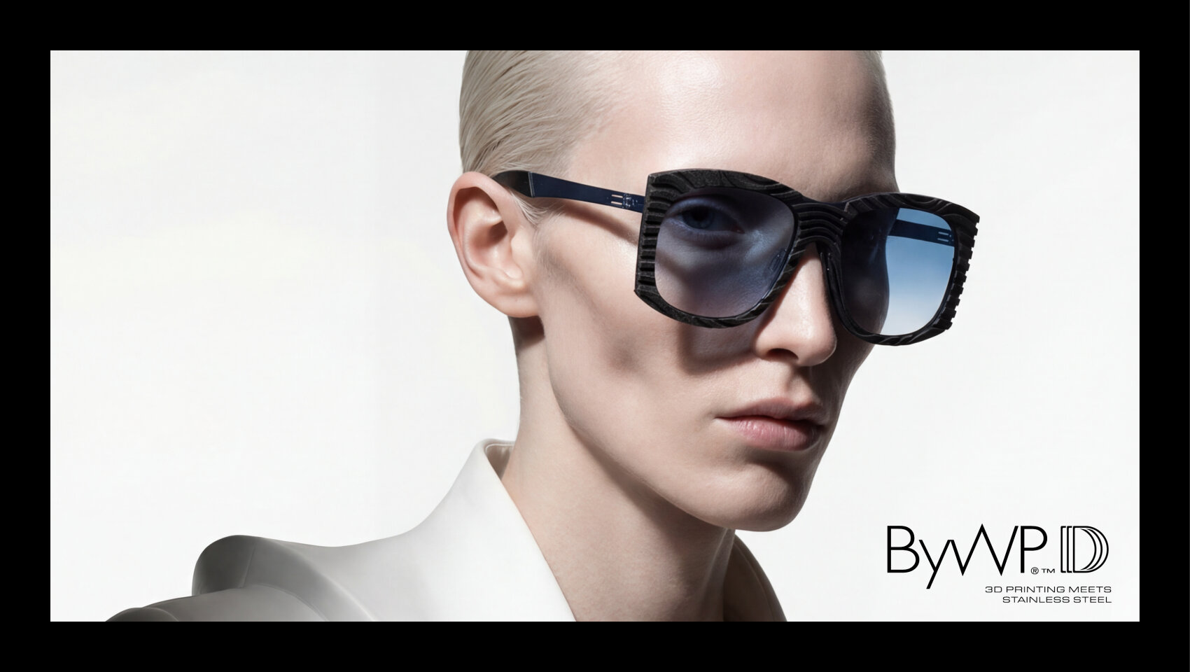 Eine Person mit kurzen, zurückgegelten blonden Haaren trägt eine große, geometrische blaue Sonnenbrille und ein weißes Hemd mit Kragen vor einem hellen Hintergrund. Unten rechts steht: ByWP: 3D-Druck trifft auf Edelstahl.