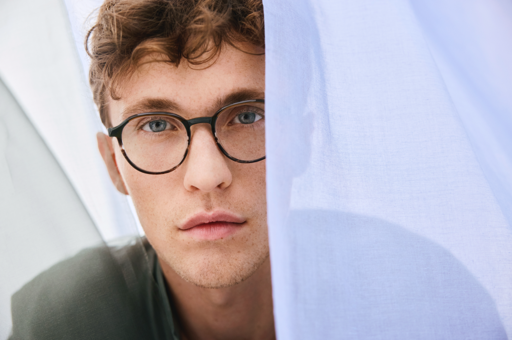 A young person with short curly hair and round glasses looks directly at the camera, partially framed by soft, sheer white fabric draped around them.