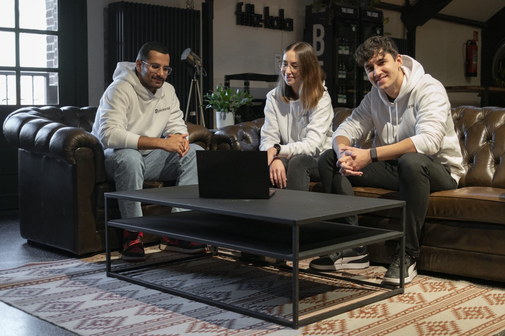 Three young adults in white hoodies sit together on brown leather sofas around a black coffee table with a laptop, smiling and chatting in a cozy, modern room with plants and warm lighting.