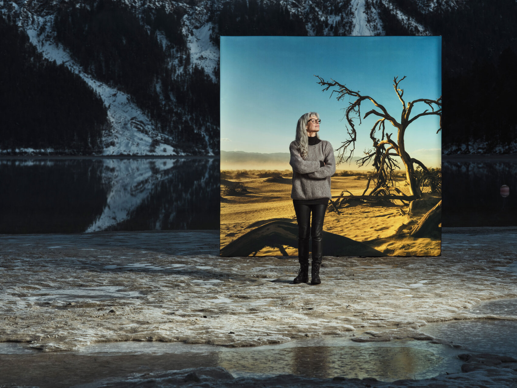 A woman in a gray sweater and black trousers stands on a snow-covered surface against the backdrop of a dry, sunlit landscape with a bare tree contrasting with snow-covered mountains and a lake in the real background.
